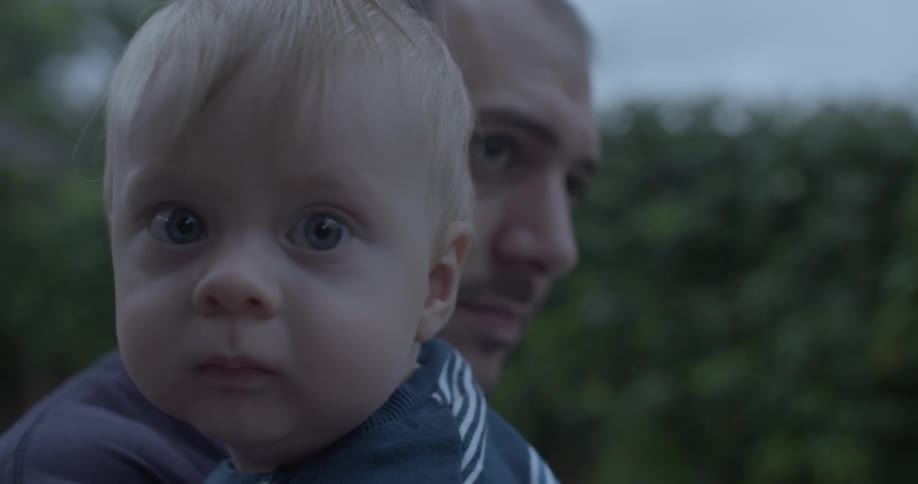A baby stares into the camera while being held by an adult man outdoors in soft natural lighting