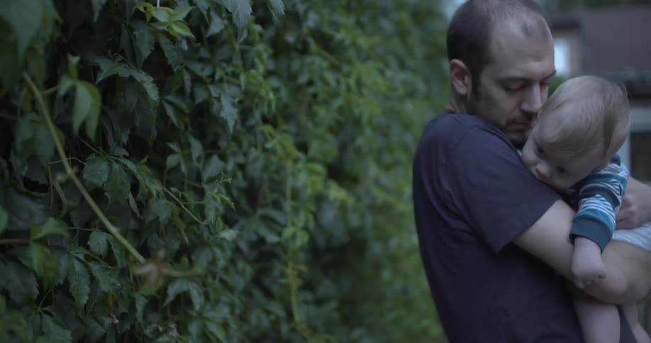 Father carries son on his chest while standing outdoors in front of a plant fence during daytime