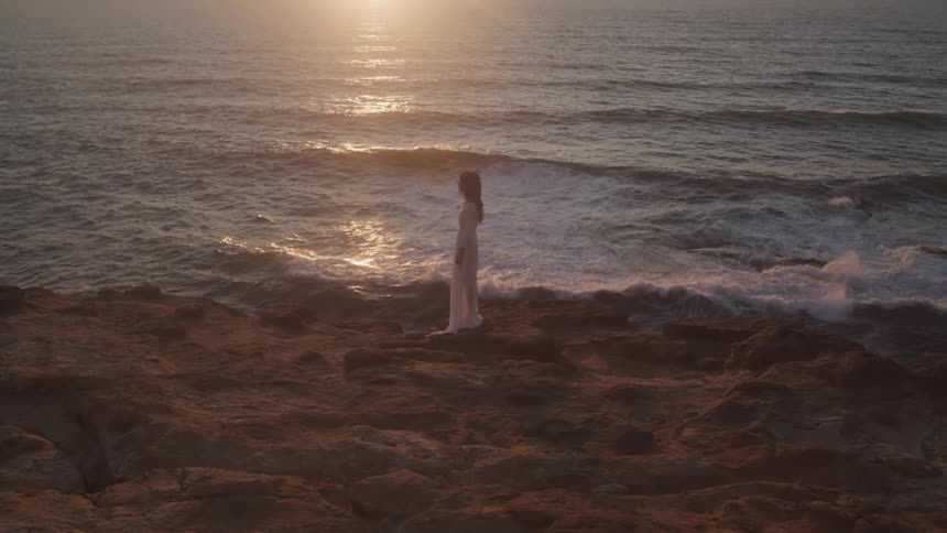 A woman stands on a beach in a white flowy dress