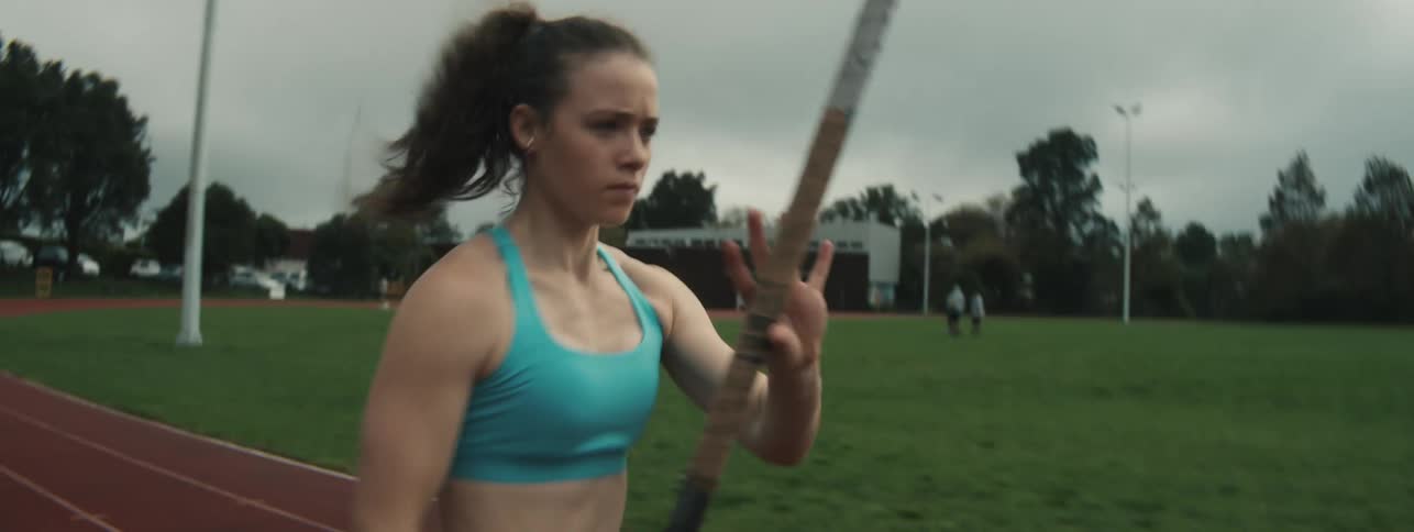 A determined female athlete runs on a track holding a pole for pole vaulting