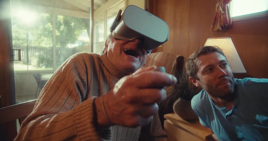 An elderly man wears a virtual reality headset while a younger man tells him what to do