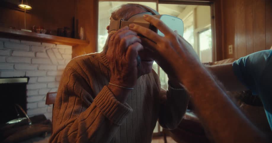 Younger man helps an elderly man try on a virtual reality headset while they sit in the living room