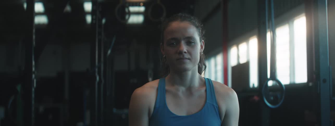 A fit young woman in athletic wear stands confidently in a dimly lit gym looking directly at the camera