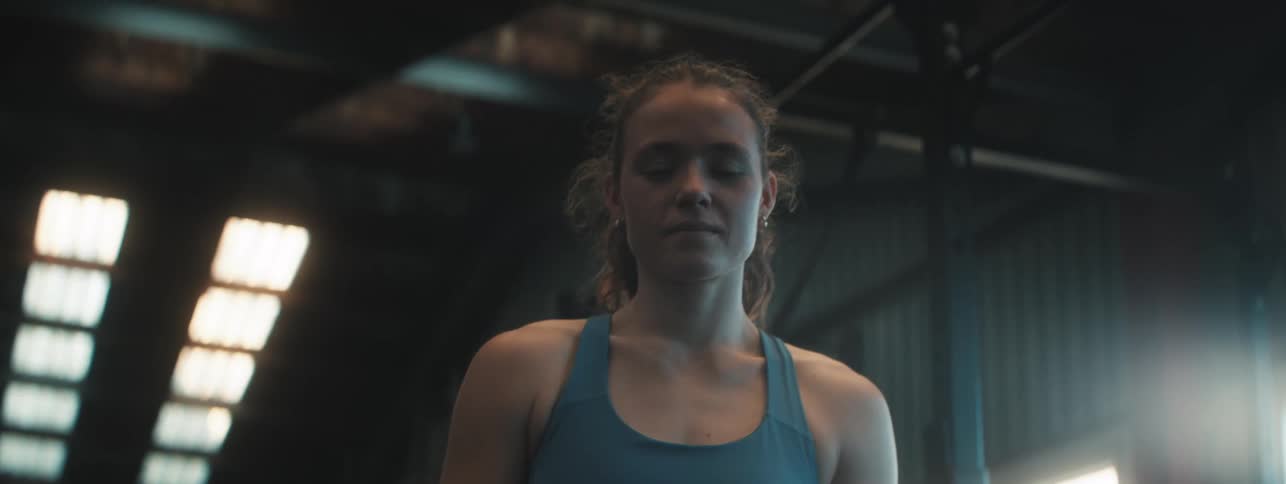 A woman in a blue tank top stands in a dim industrial gym surrounded by metal beams