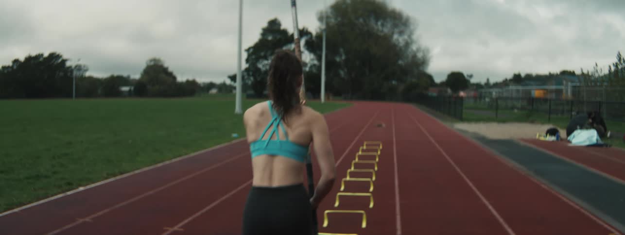 A woman in athletic wear stands on a running track preparing for pole vault practice with a row of evenly spaced yellow hurdles ahead