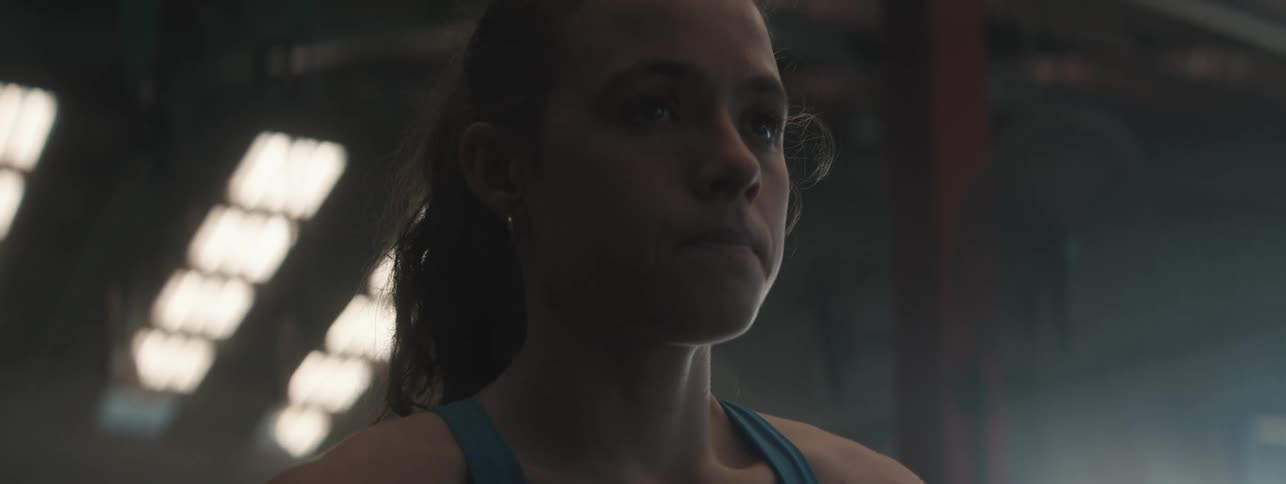 A woman wearing a blue tank top catches her breath in a dimly lit room with bright overhead lights