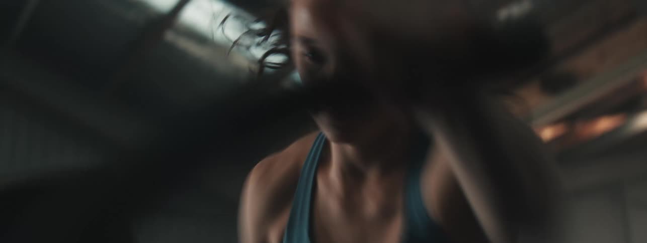 A female athlete in a blue sports bra powers through an intense elliptical workout in a dimly lit gym with a corrugated metal ceiling and industrial fan overhead