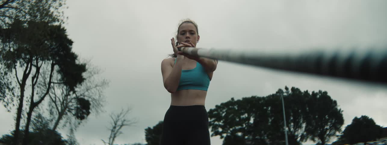 A focused female pole vaulter in a blue sports bra and black leggings firmly grips a pole while looking forward under an overcast sky with trees in the background