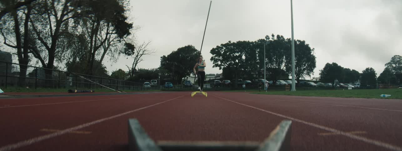 Female pole vaulter trains by running over very low hurdles while others walk on the running track on a summer day