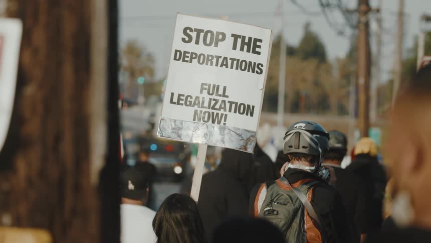 A diverse crowd of protesters gathers on a street some holding signs facing several white armored vehicles and police cars under an overcast sky