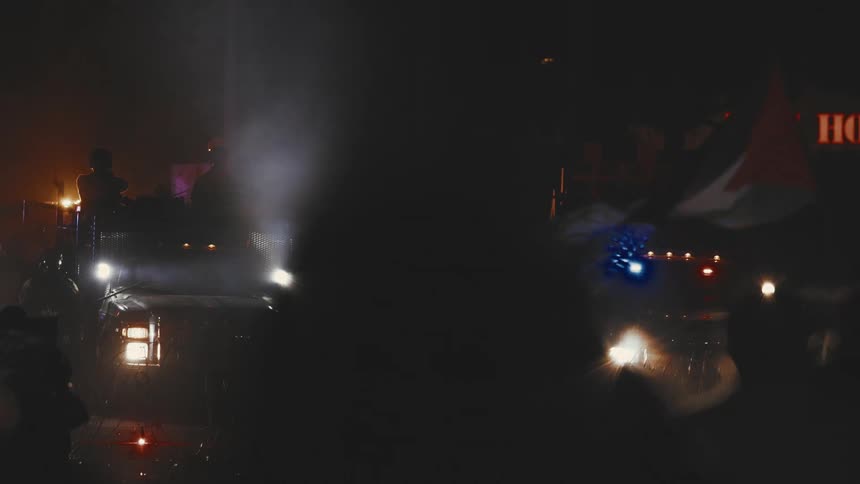 A diverse crowd of protesters gathers on a street at night some holding flags facing several armored vehicles and police cars
