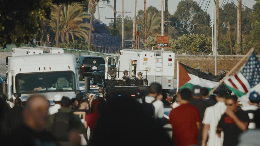 A diverse crowd of protesters gathers on a street some holding flags facing several white armored vehicles and police cars under an overcast sky