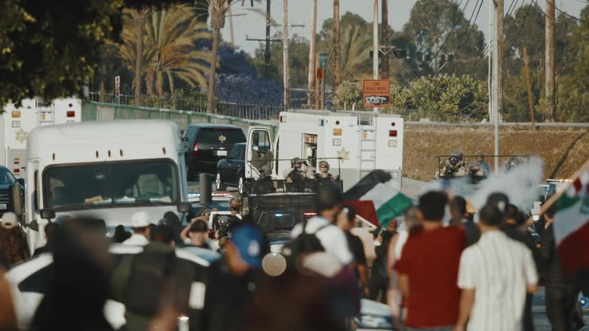A diverse crowd of protesters gathers on a street some holding flags facing several white armored vehicles and police cars under an overcast sky