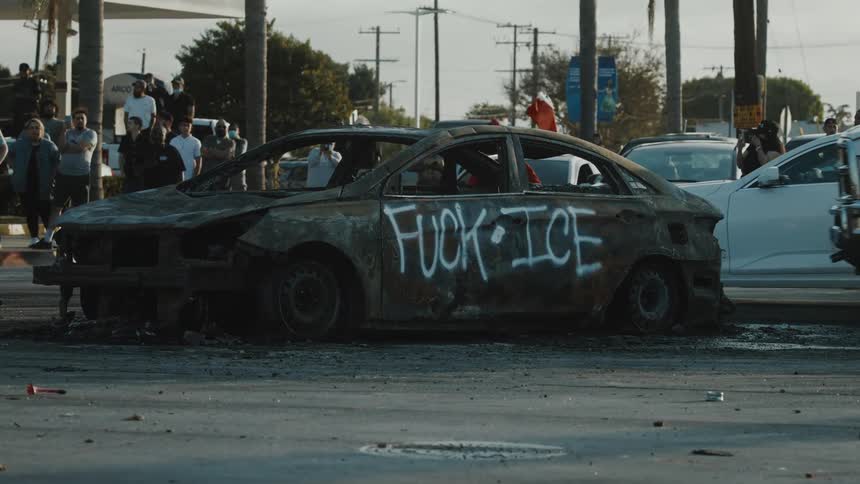 Traffic passes a car carcass protesting the ice agency while a camera operator films and people stand nearby