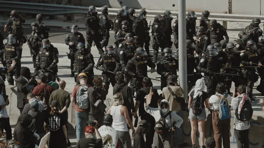 A tense confrontation between a large group of riot police in tactical gear and a crowd of civilians in an urban setting
