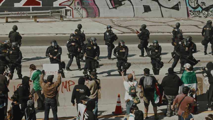 A tense confrontation between a large group of riot police in tactical gear and a crowd of civilians in an urban setting