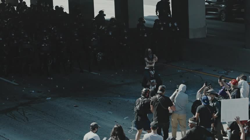A group of people stands under an overpass bridge during a protest holding placards and signs as they face off with policemen