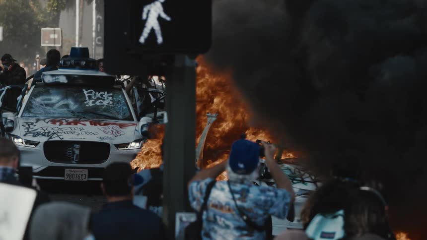 A car burns during a street riot at midday as journalists take photos of the scene