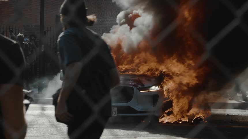 A white car with flames and smoke erupting from its front is parked on a street with people standing nearby and a chainlink fence in the foreground