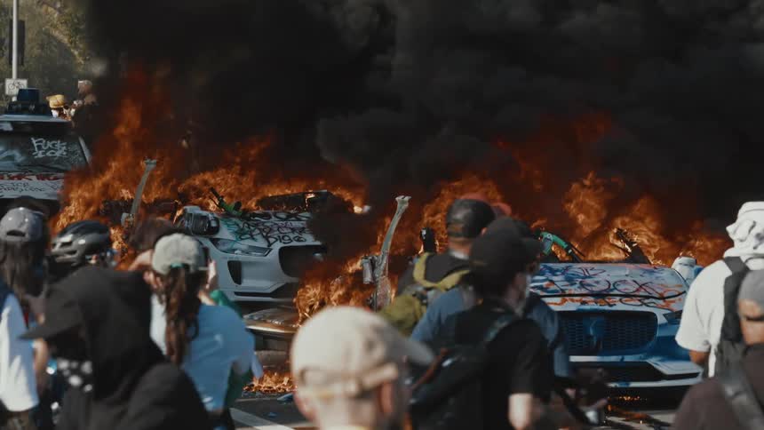 The cars are burning as the rioters walk around the road