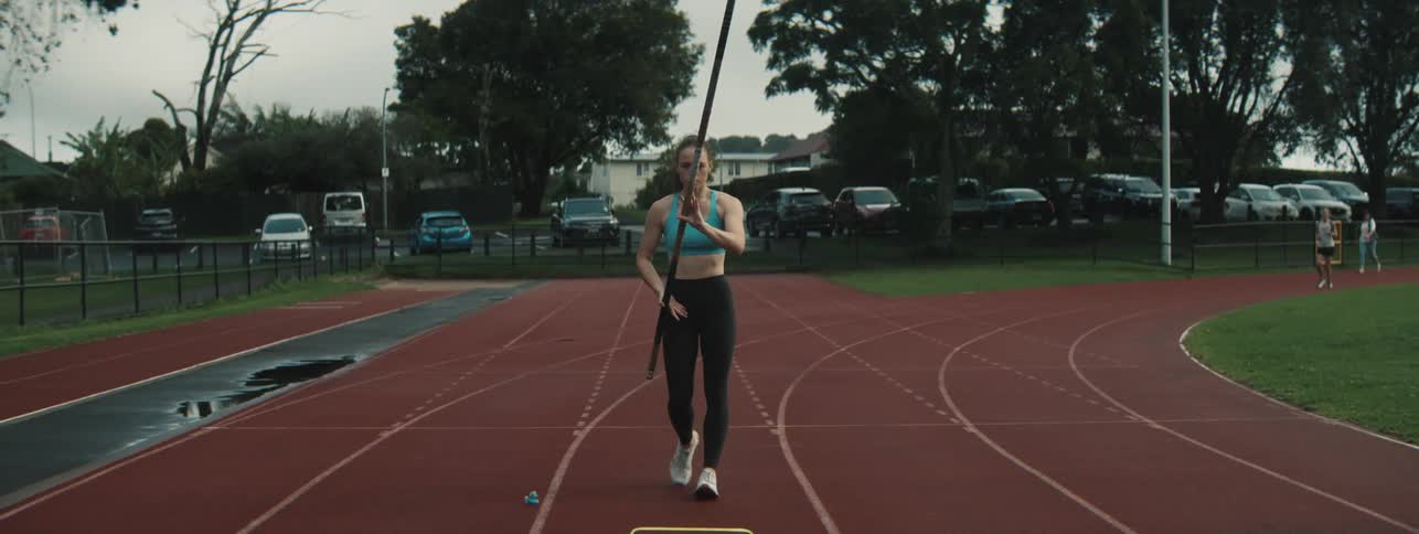 A female athlete runs on a red track holding a pole under a cloudy sky