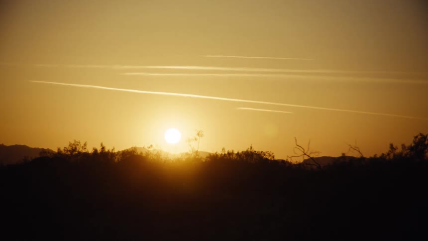 A large bright sun sets behind a dark silhouetted line of low hills and sparse vegetation with several prominent contrails streaking across the yellow and orange sky