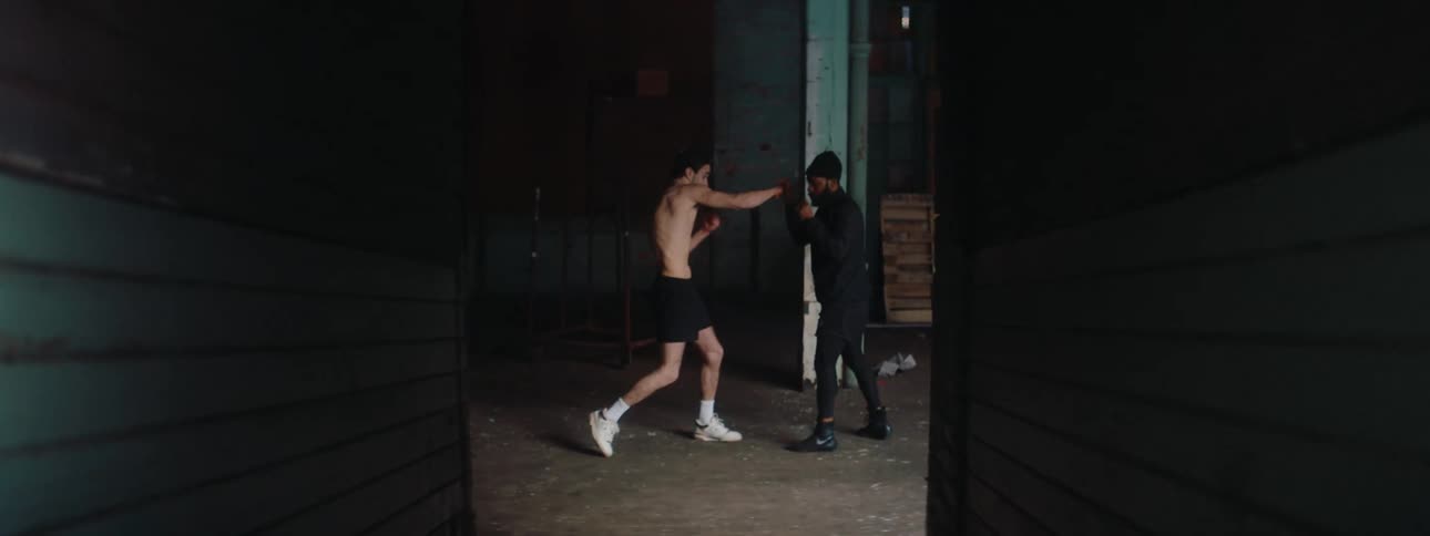 A shirtless man is boxing with a person standing in front of him in an indoor setting