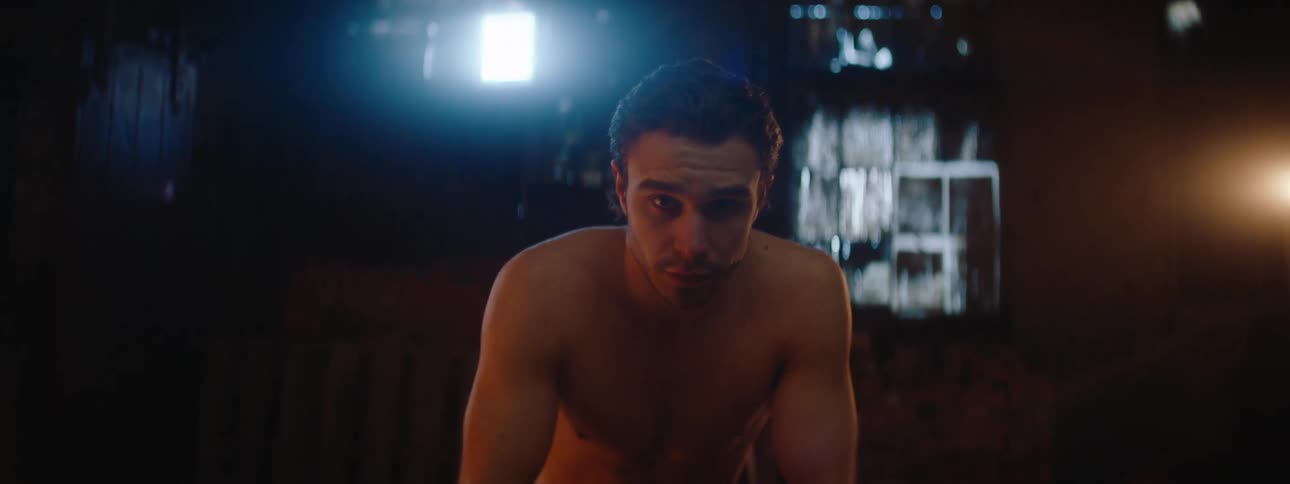 A shirtless person sits on a chair in a dimly lit room with wooden pallets and a side light casting shadows across the scene