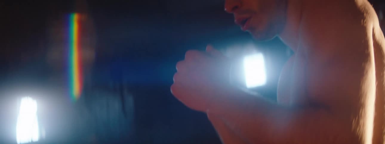 A shirtless man is throwing punches in front of him and there are some lights behind him