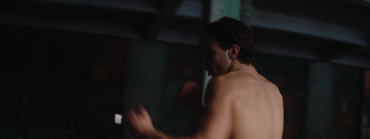 A shirtless man trains alone in a dim empty warehouse with exposed brick walls and wooden paneling