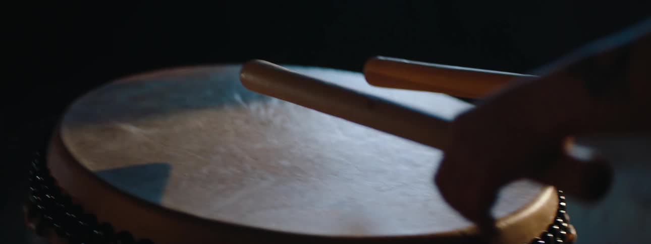 A persons hands holding drumsticks and striking a hand drum with dynamic lens flares and light effects throughout the shot