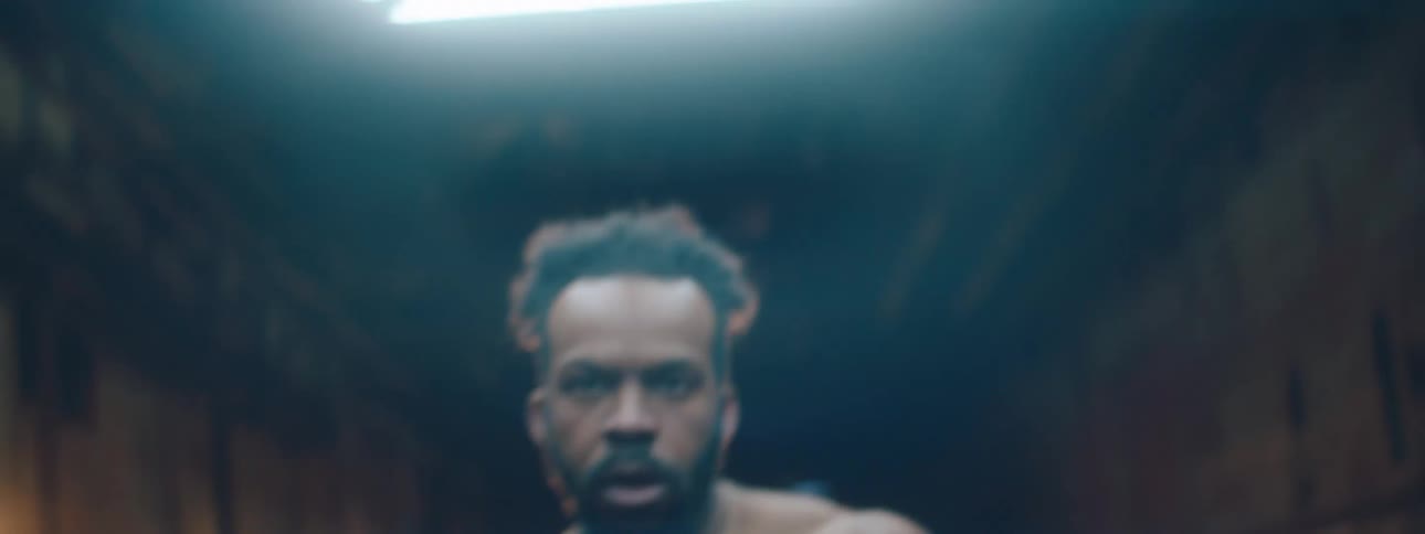 A muscular shirtless man with a beard is shown from the waist up boxing on a dark street at night with blurred lights in the background
