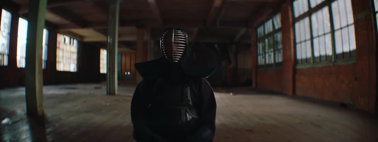 A kendo practitioner wearing a helmet and black uniform sits on the floor of a wide empty room holding a sword calmly across their lap