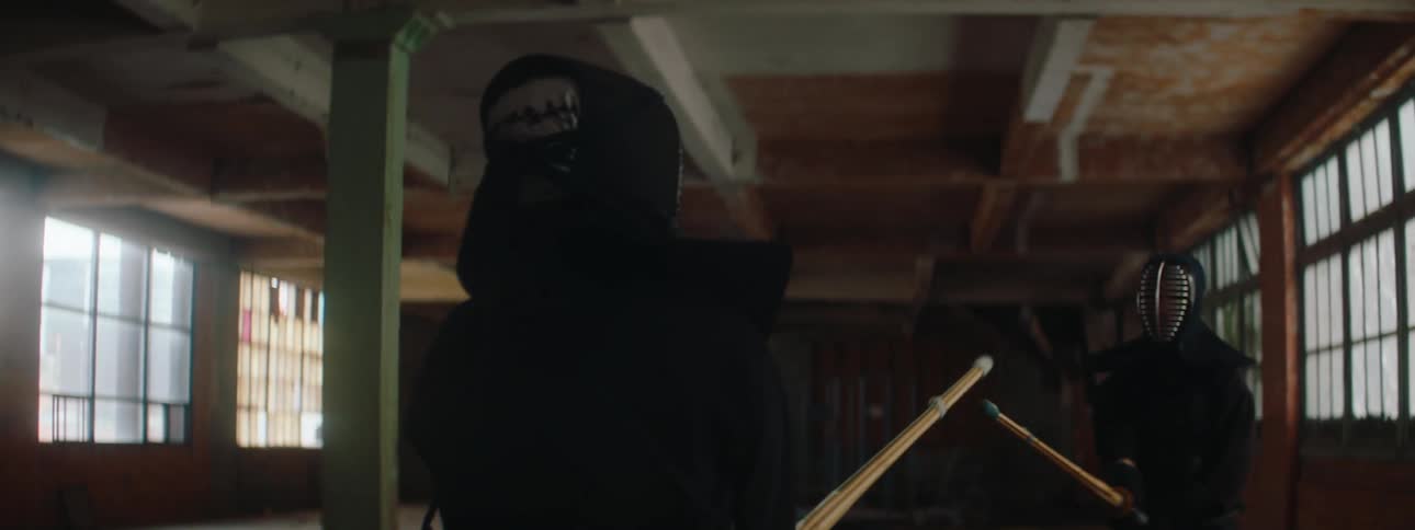 Two people in kendo armor spar with bamboo swords in a dim room with large windows