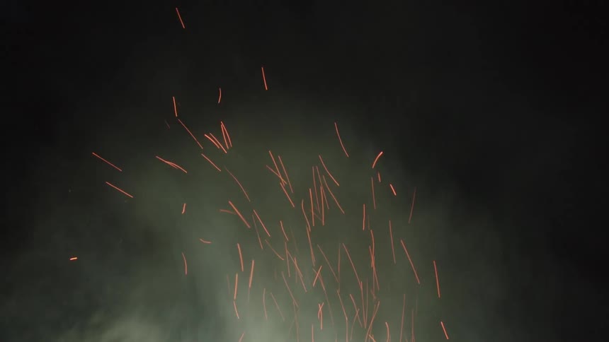 Numerous orange sparks and light with smoke billow rising upwards against a dark background