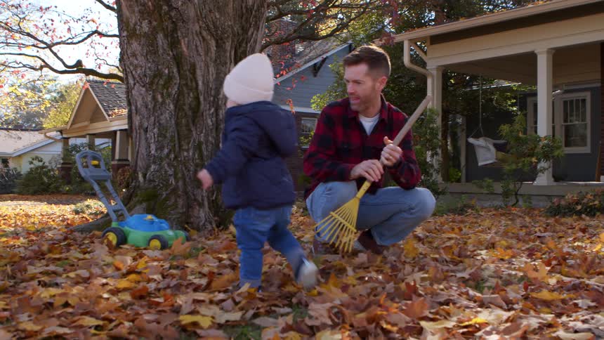 Baby walks through autumn leaves while a smiling man kneels nearby with a rake