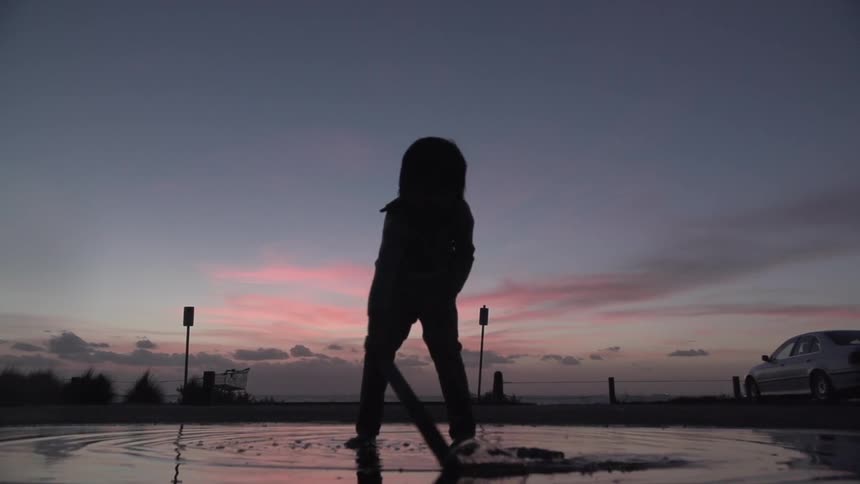 A child plays in a puddle with a stick at dusk
