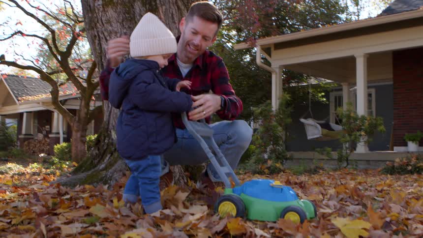A father helps his toddler play with a toy lawnmower in a leafcovered yard during fall
