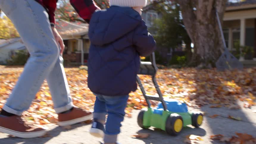 Father guides son as he plays with a toy lawn mower on a pathway outdoors during daytime