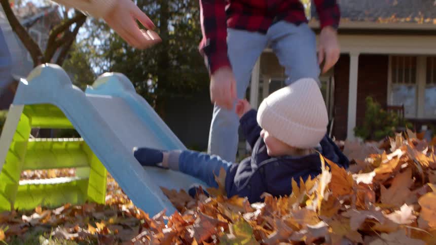 A father helps his toddler slide into autumn leaves as the mother playfully catches them