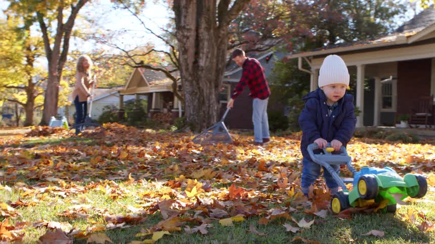 A child plays with a toy lawn mower in the front yard at midday while the parents rake fallen leaves scattered across the ground