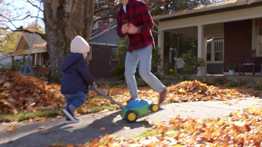 A father takes a picture of his toddler pushing a toy lawnmower outside in the fall