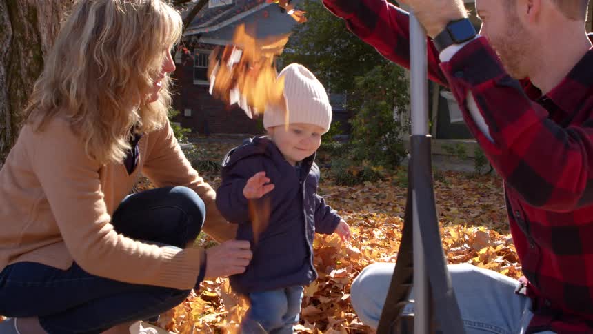 A family pauses from raking to play in autumn leaves outside their house on a sunny day