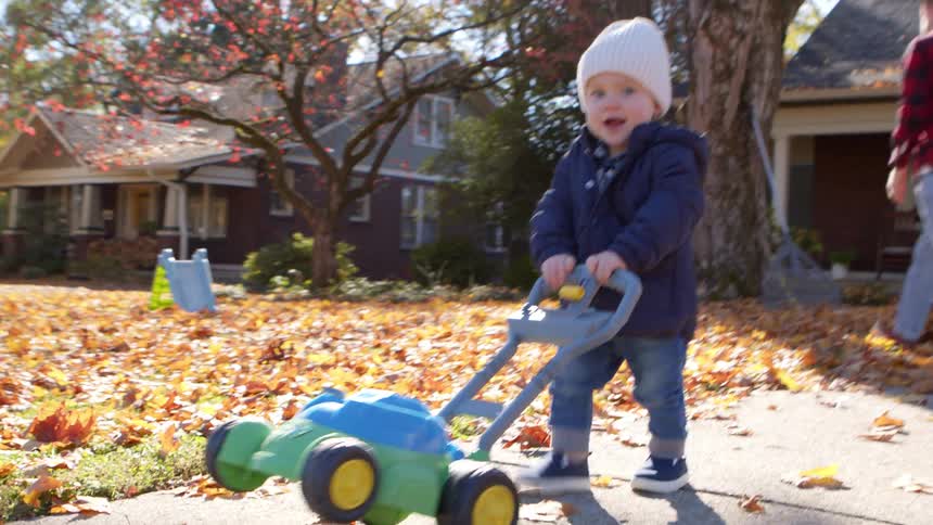 A toddler pushes a lawnmower toy outside in the fall while his father watches him