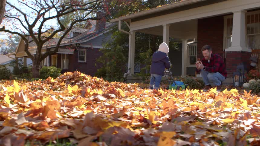 Father records son playing with a toy lawn mower outdoors in front of their home during daytime