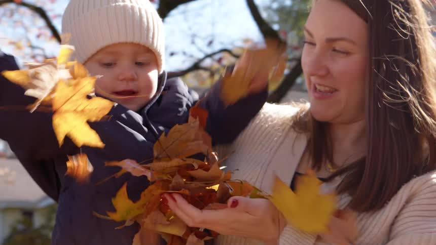 A mother holds her child who is dressed in a winter hat and jacket as the child receives leaves from another person and joyfully scatters them on the ground while laughing at midday