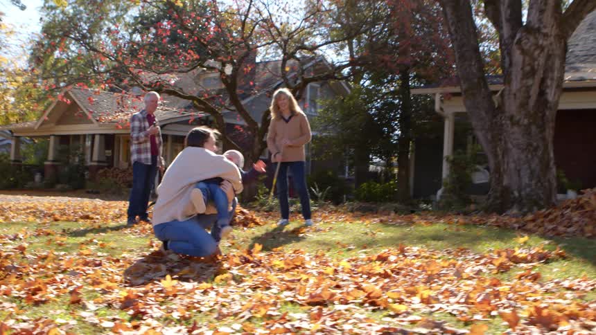 A woman kneels in a leafstrewn yard playing with a child while other adults linger in the background amid houses and autumncolored trees