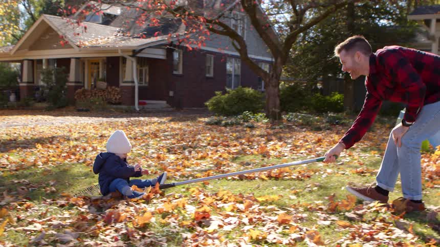 A father playfully pulls his seated son with a rope in a leafy yard while the mother assists from behind