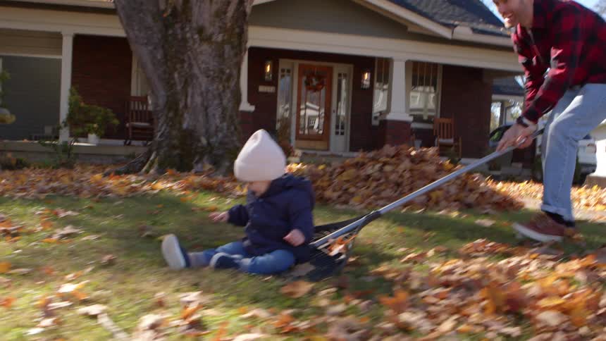 A man pulls a toddler on a tarp through autumn leaves as a woman watches