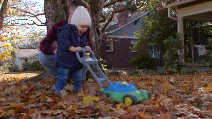 A father helps his toddler son push a toy lawnmower in the leafcovered lawn in the fall